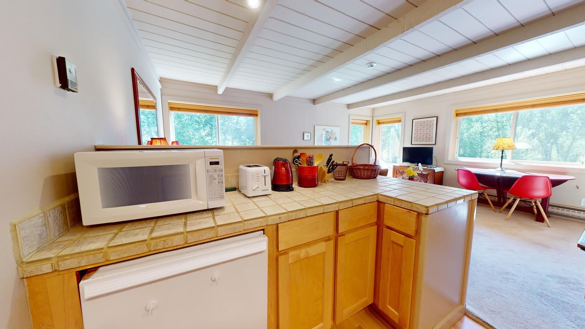 A kitchen with a microwave, toaster, and red kettle on the counter, wooden cabinets, and a view of the outdoors through large windows.