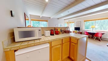 A kitchen with a microwave, toaster, and red kettle on the counter, wooden cabinets, and a view of the outdoors through large windows.