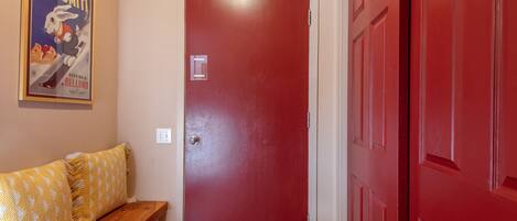 A red door with a small window, a wooden bench with yellow patterned cushions, and a poster on the wall.