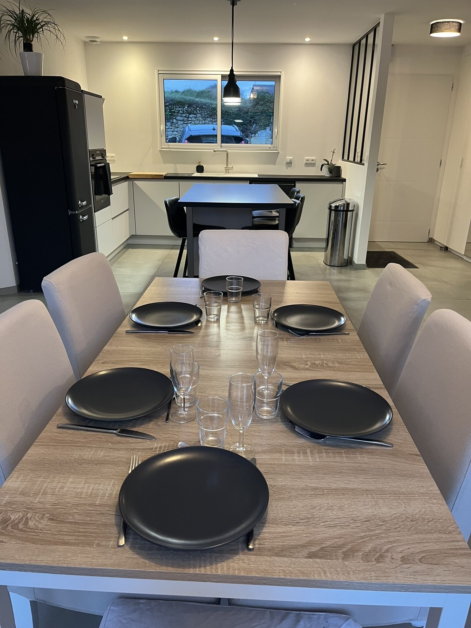Een moderne eethoek met een lange tafel gedekt voor vier personen, een zwarte koelkast, en een raam met uitzicht op een stenen muur en begroeiing.