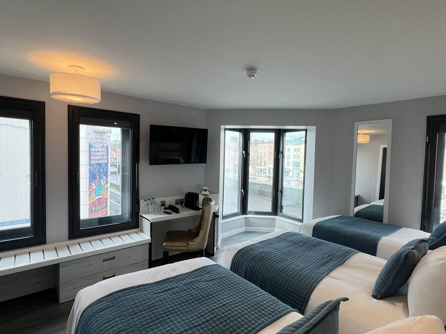 Habitación de hotel con dos camas, un escritorio, una silla, un televisor y ventanas amplias con vistas a la ciudad.