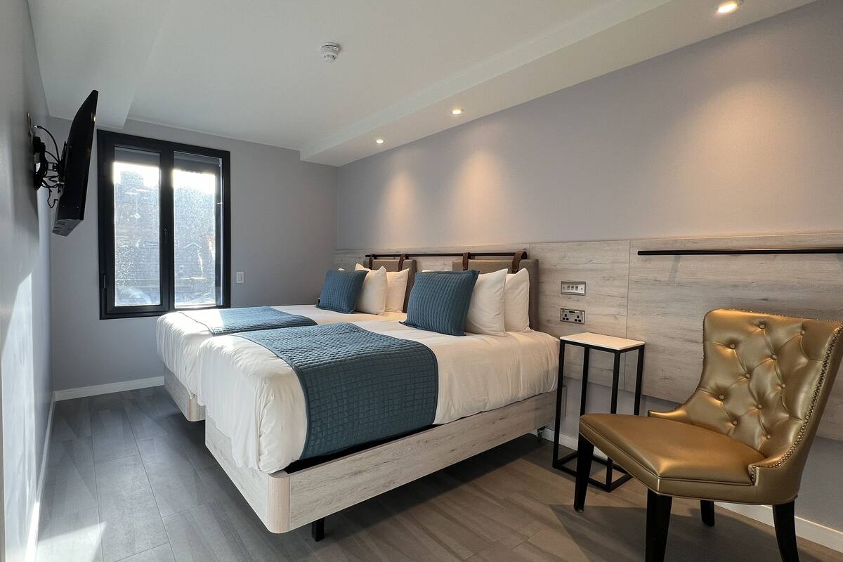 A modern bedroom with a large bed, a side table, a gold chair, and a window with a view of the city.