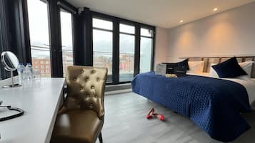 A hotel room with a bed, a chair, a desk with a mirror, and a view of buildings through large windows.