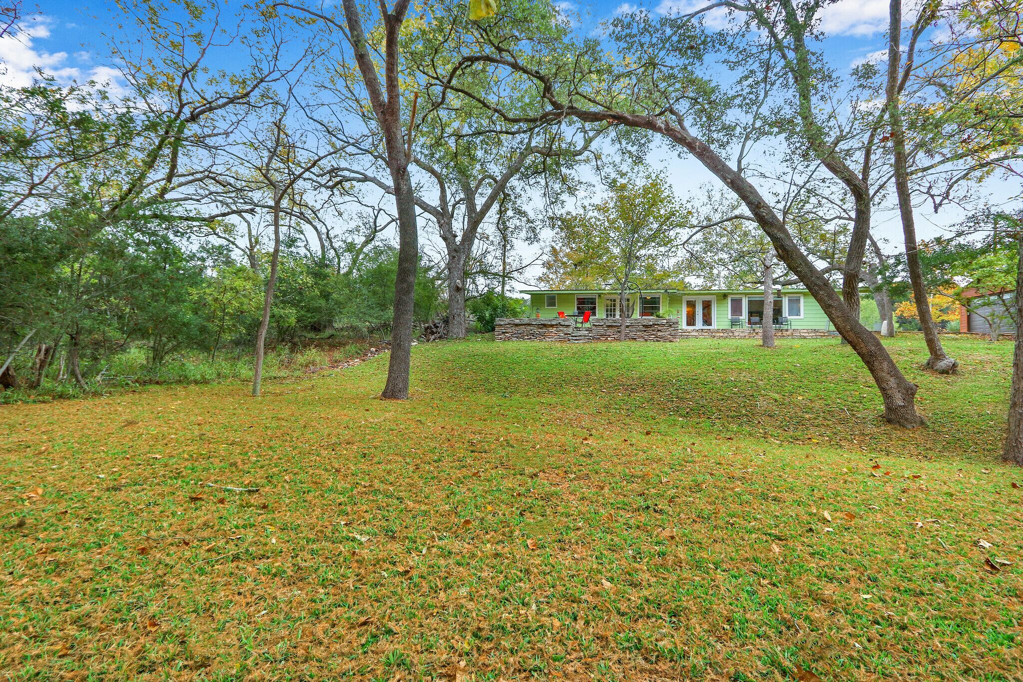 A green house with a stone foundation, surrounded by trees and a grassy yard.
