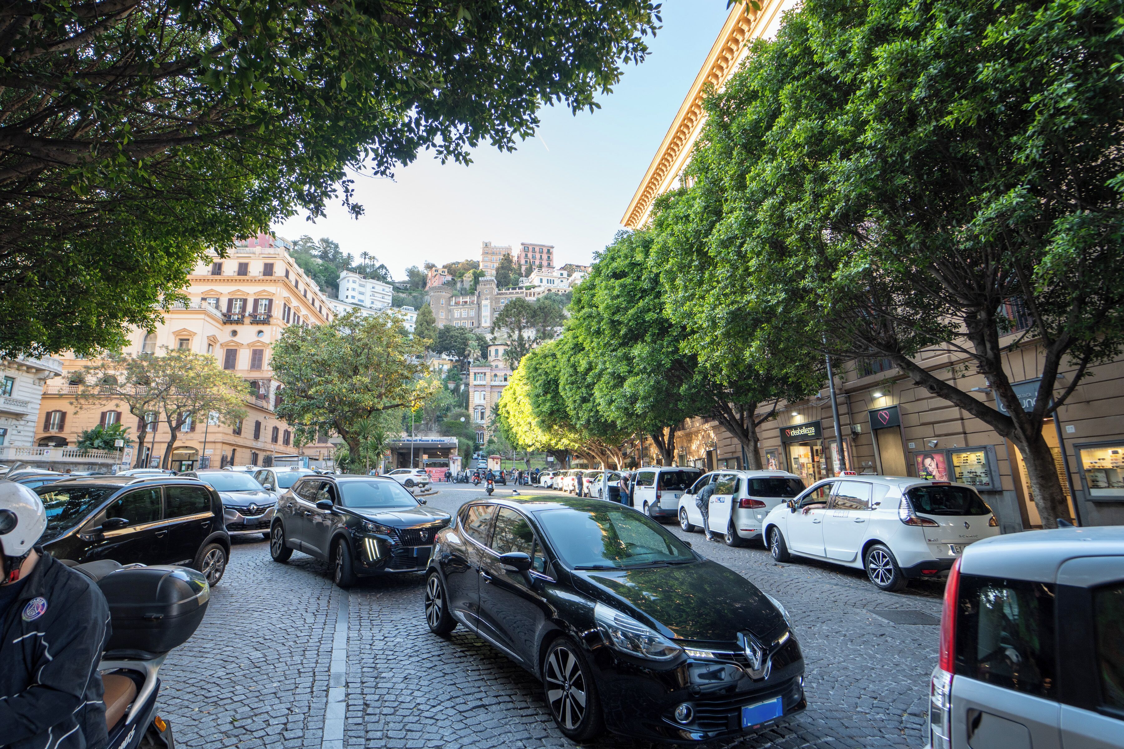 Une rue pavée bordée de voitures garées et d’arbres, qui mène à un immeuble à plusieurs étages doté de balcons.
