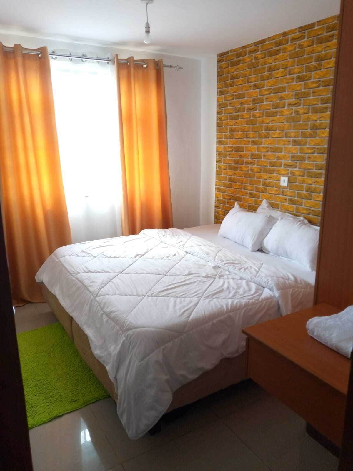 Una cama bien tendida con ropa blanca en una habitación con pared de ladrillo y alfombra verde.