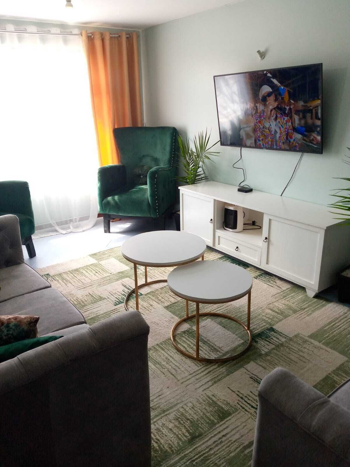 Een woonkamer met een groene fauteuil, twee ronde tafels, een witte kast en een aan de muur bevestigde televisie.