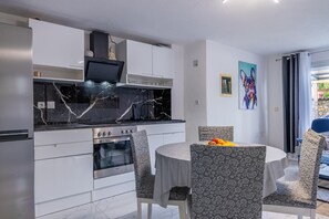 Eine moderne Küche mit weißen Schränken, einer schwarzen Marmorarbeitsplatte und einem eingebauten Backofen. Ein Esstisch mit gemusterten Stühlen und ein farbenfrohes Gemälde an der Wand sind ebenfalls vorhanden.