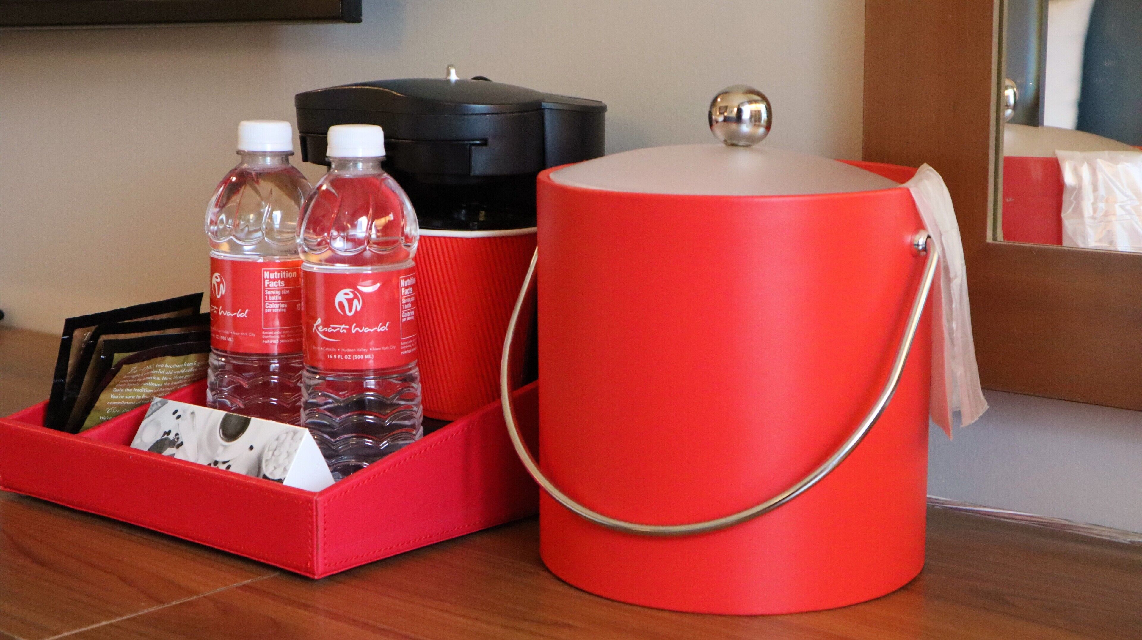 Un mini-bar rouge contenant une bouteille d’eau, une bouteille de jus de fruits et un récipient noir, qui pourrait être une machine à café, le tout posé sur une surface en bois.