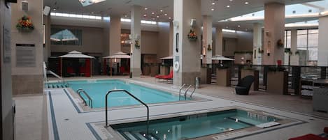 A large indoor swimming pool with multiple lanes, surrounded by lounge areas with red chairs and umbrellas, and a high ceiling with recessed lighting.