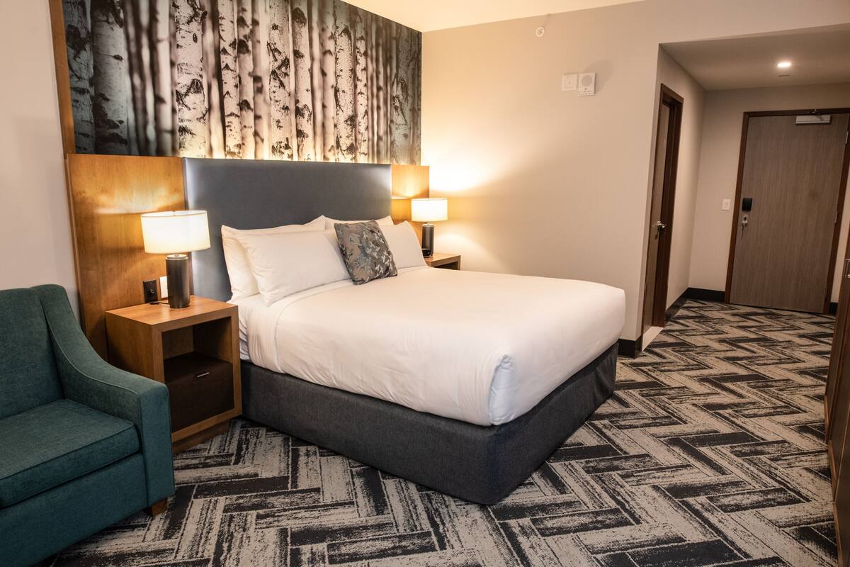 Een hotelkamer met een groot bed, twee nachtkastjes met lampen, een blauwgroene fauteuil en een kamerbreed tapijt met dessin.