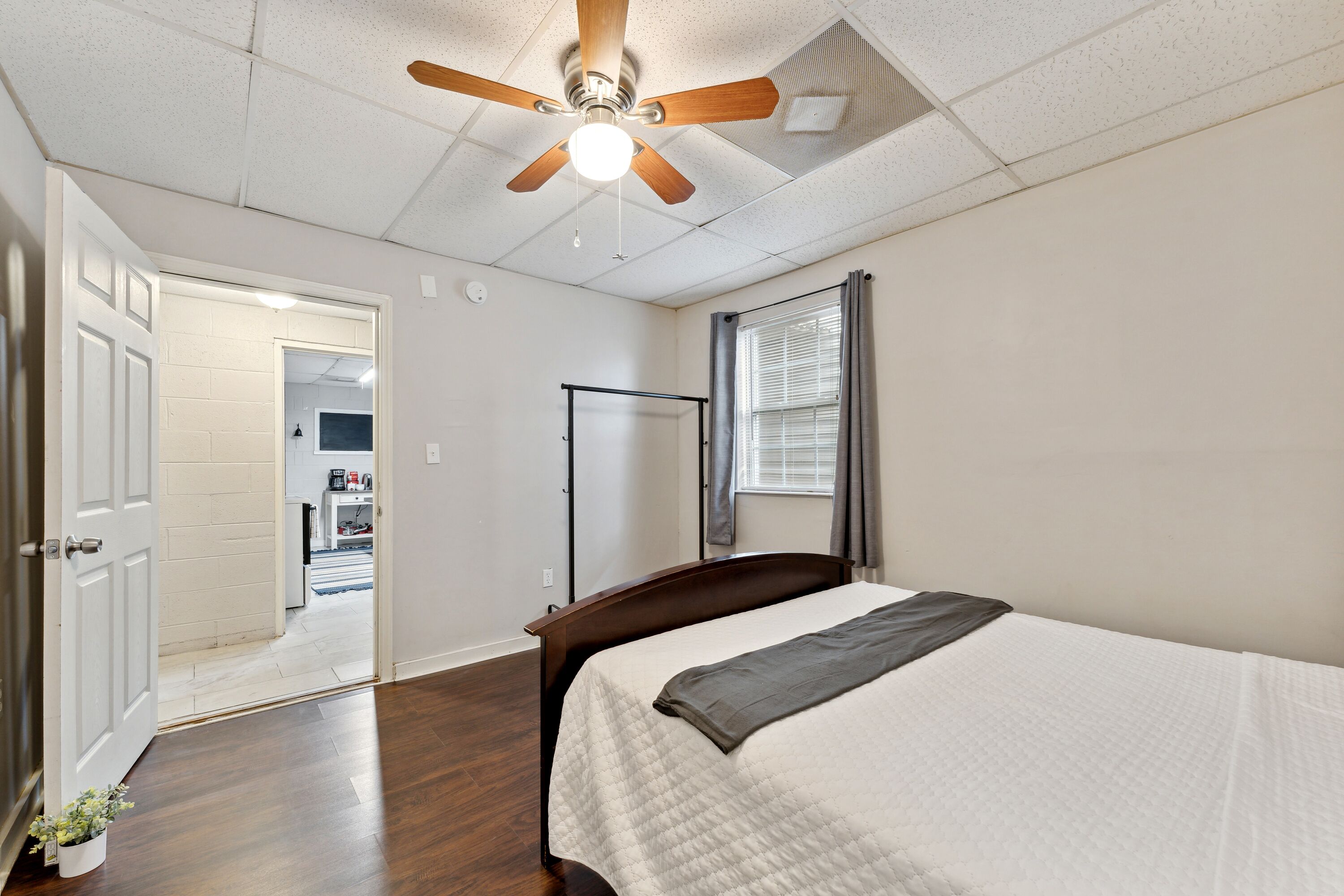 A bedroom with a bed, a ceiling fan, a window with curtains, and a door leading to a hallway.