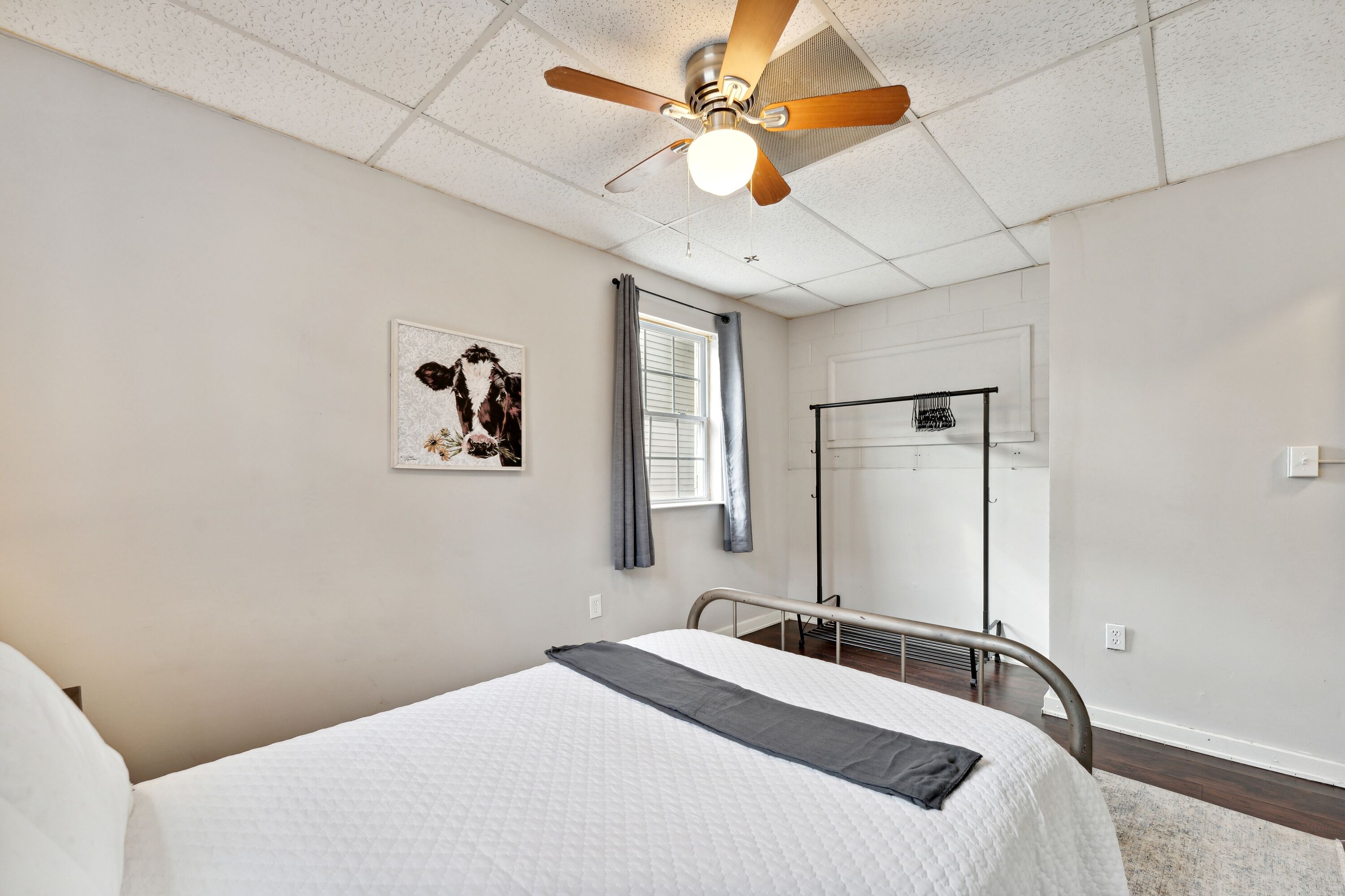 Ein Schlafzimmer mit einem Bett, einem Deckenventilator, einem Fenster mit Vorhängen, einer Wandheizung und einem gerahmten Bild.