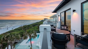 A balcony with a pool, outdoor seating, and a view of the beach at sunset.