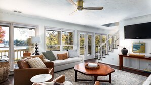 A spacious living room with a large window, a ceiling fan, a sofa, a coffee table, and a television. - Beautiful Private Home in Quiet Cove. Swim Dock, Kayaks, Stand Ups.  Boat Option (York)