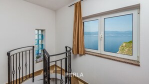 Una habitación con un ventanal grande con vistas al mar, un ventanuilla con un marco decorativo, y una escalera con barandilla de metal.