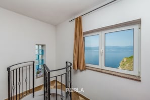 A room with a large window overlooking a coastal view, a small window with a decorative frame, and a staircase with a metal railing.