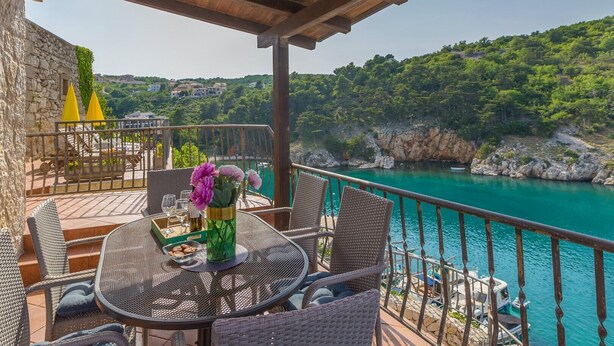 Una terraza con mobiliario de mimbre, una mesa con un jarrón de flores y vistas al mar turquesa y colinas verdes.