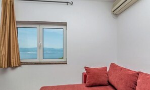 A room with a bed, a window with a view of the sea, and an air conditioning unit.