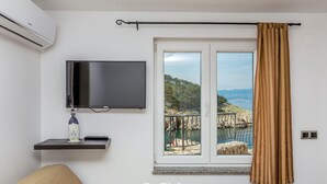 Una habitaci ón con televisión, un estante pequeño con un objeto decorativo, una ventana con vista al mar y una cortina.