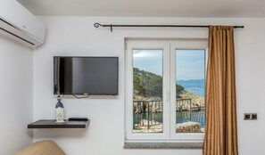 A room with a TV, a small shelf with a decorative item, a window with a view of the sea, and a curtain.