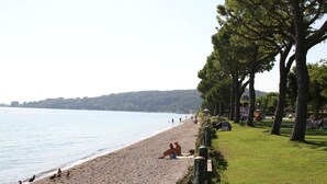 Ein Strand mit Erholungssuchenden, eine begrünte Fläche mit Bäumen und ruhigem Wasser.