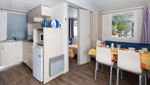A compact kitchen with a refrigerator, a microwave, and a sink. There is a dining area with a table and chairs, and a window with curtains. - Ferienpark Cesenatico Mobilehome Riviera für 6 Personen (Cesenatico)