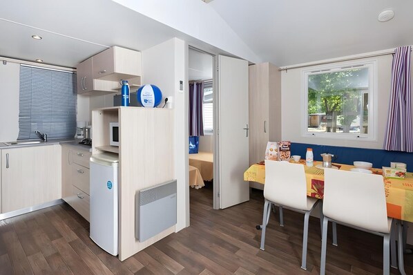A compact kitchen with a refrigerator, a microwave, and a sink. There is a dining area with a table and chairs, and a window with curtains. - Ferienpark Cesenatico Mobilehome Riviera für 6 Personen (Cesenatico)