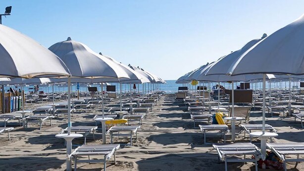 Ein Strand mit Reihen von Liegestühlen und Sonnenschirmen, einige mit Personen, und freier Blick auf das Meer.