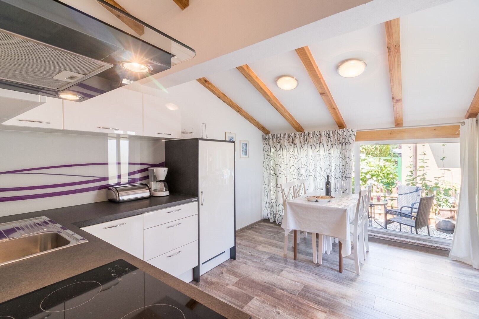 Cozinha moderna com armários brancos, forno embutido e cafeteira. Há uma área de jantar com mesa e cadeiras, e vista para o exterior através de uma janela com cortinas.