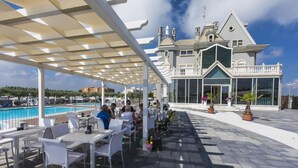 An outdoor patio with white tables and chairs, a pool, and a large building with a balcony in the background.