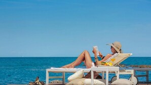 Una persona descansando en una tumbona junto al mar, con un libro y una bebida sobre la mesa.