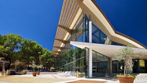 Un edificio moderno con un diseño de techo inclinado distintivo, amplios ventanales de vidrio y un área exterior pavimentada con asientos y jardineras.