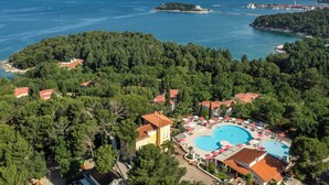 An aerial view of a coastal resort with a large swimming pool, surrounded by lush greenery and a clear view of the sea.
