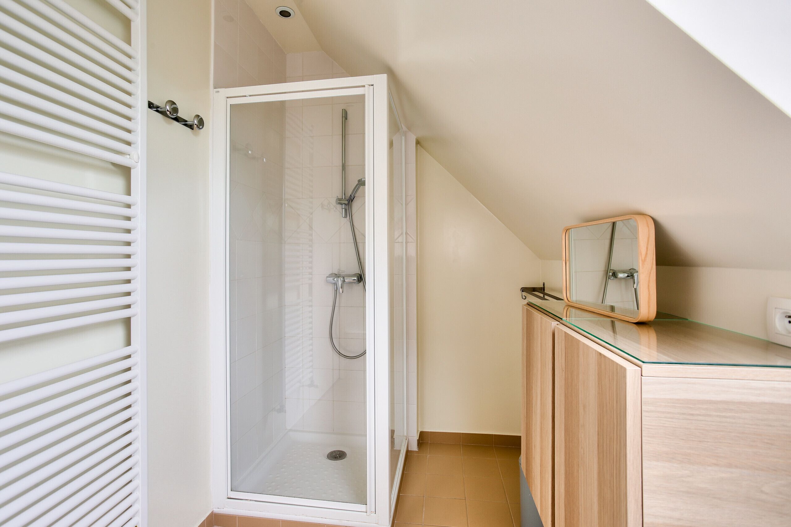 Une douche avec une porte en verre, un meuble en bois avec un miroir et un radiateur blanc.