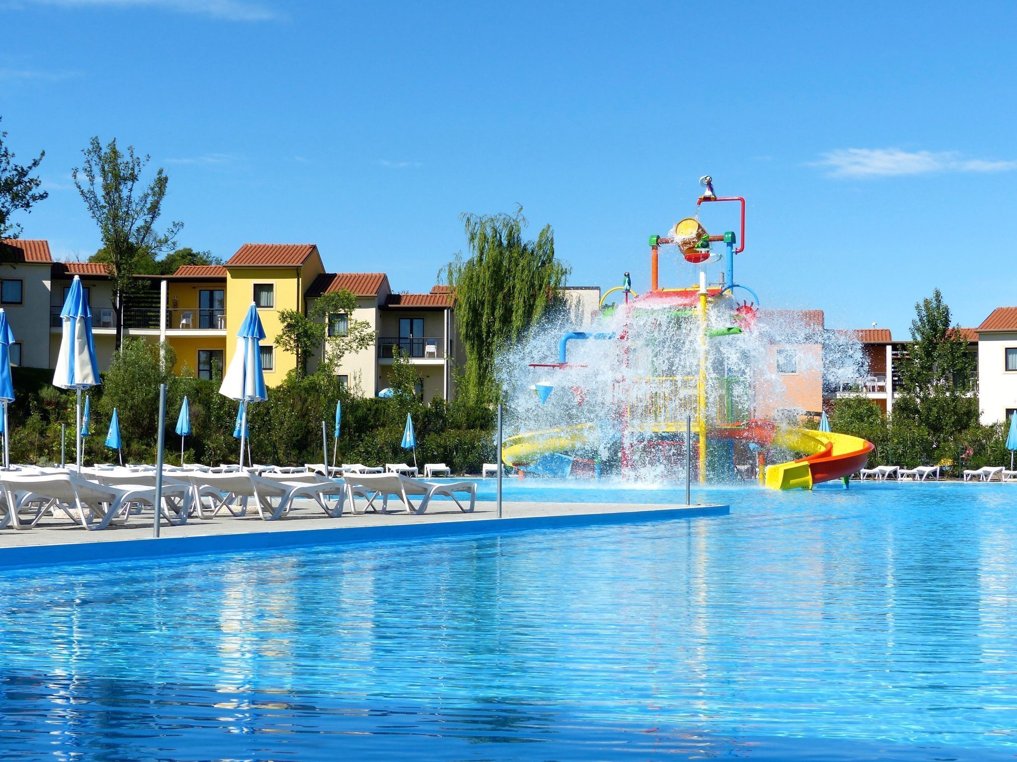 Una piscina con zona de juegos acuáticos, sillas de descanso y edificios de hotel al fondo.