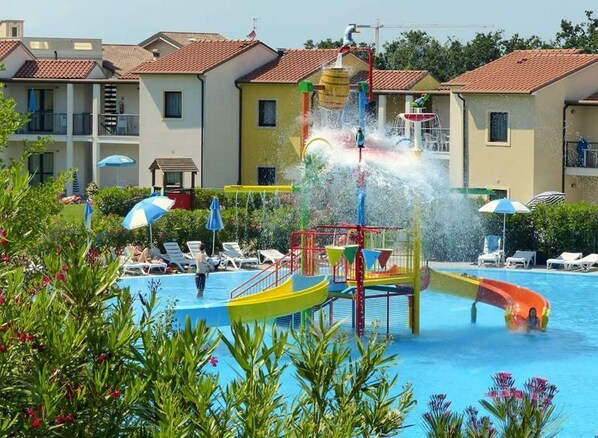 Een zwembad met waterglijbanen en een speelgedeelte, omringd door ligbedden en parasols.