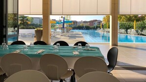 Un área de comedor al aire libre con vista a una piscina, mesas dispuestas con vasos y cubiertos, y sillas ubicadas alrededor de las mesas.