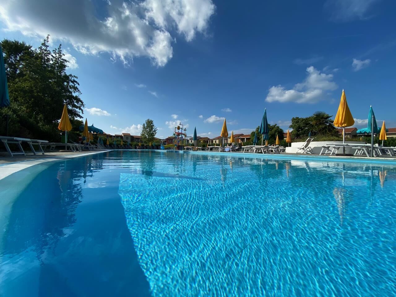 A large, clear swimming pool with lounge chairs and umbrellas, surrounded by buildings and trees.