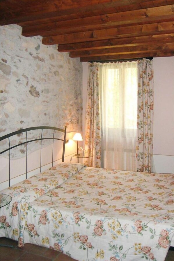 Un dormitorio con pared de piedra, techo de madera, cama con ropa de cama floral, mesita de noche con lámpara y ventana con cortinas florales.