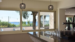 A modern kitchen with large windows, a marble countertop, and a view of the sea.