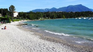 Una playa de guijarros con agua cristalina, un muelle y montañas al fondo.