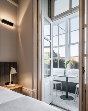 Quarto com cama, mesa de cabeceira, luminária e vista ampla para uma área externa com mobiliário.