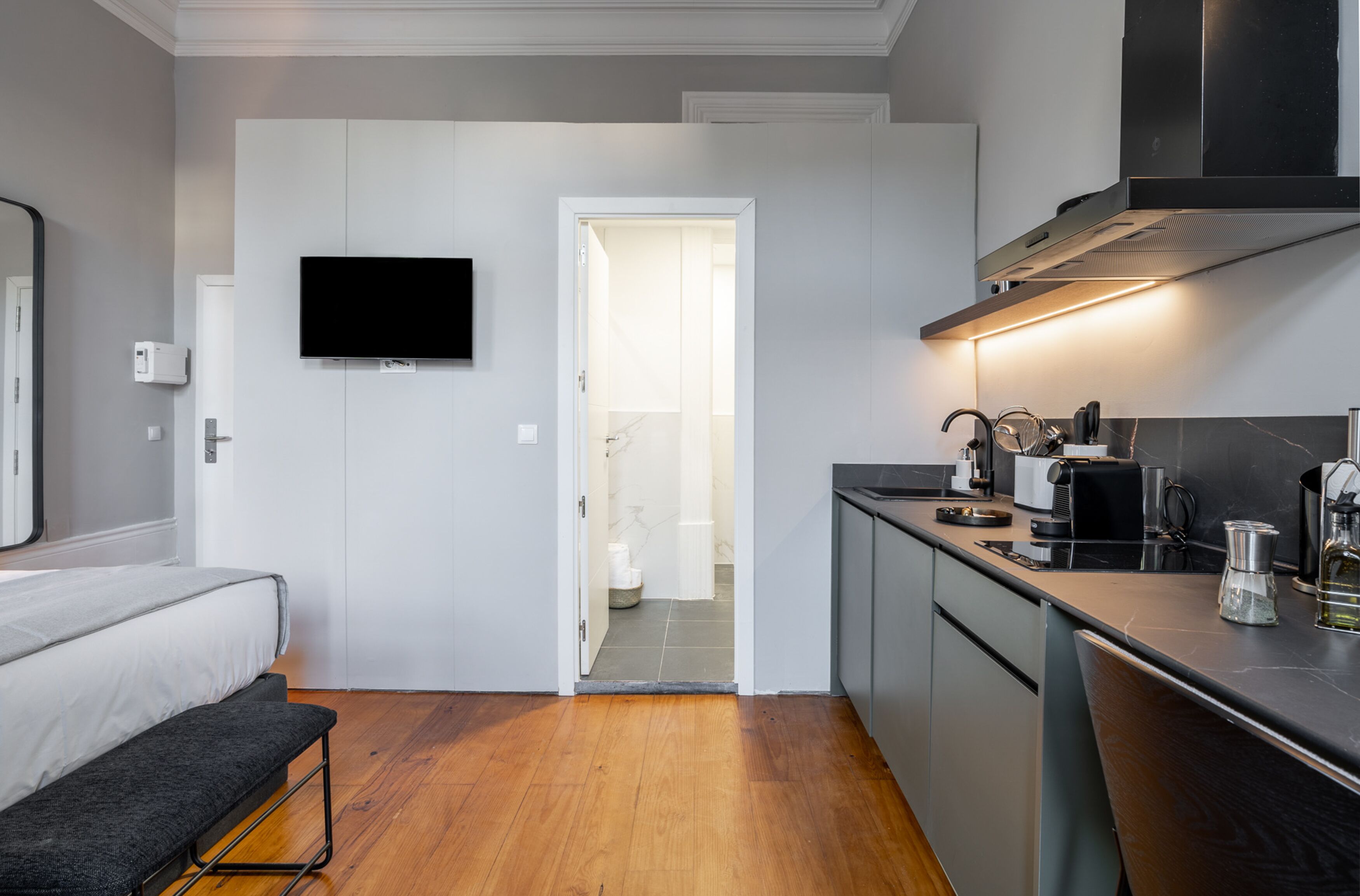 Cocina compacta con estufa integrada, microondas y cafetera. Un banco se encuentra cerca de la entrada, y un televisor está montado en la pared.