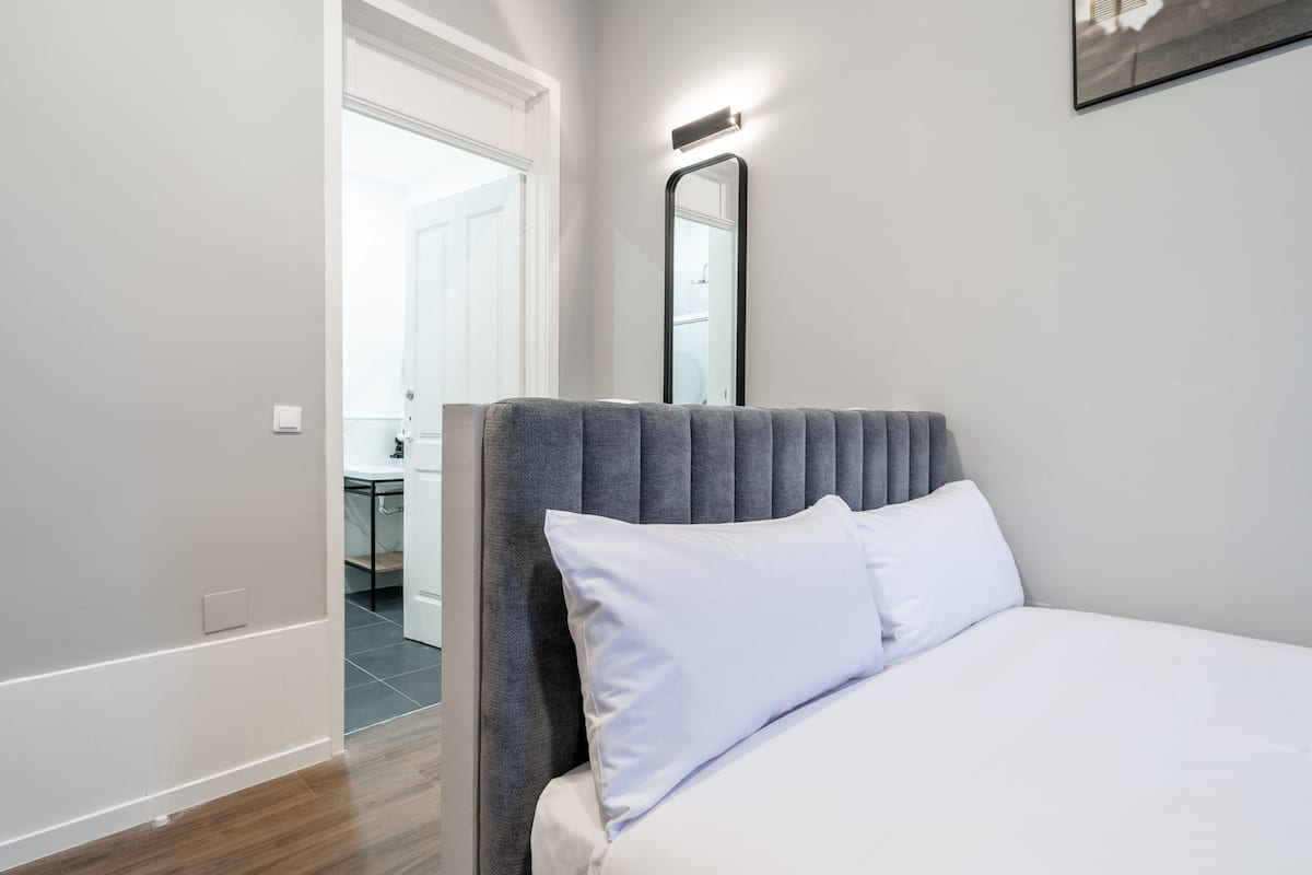 A bedroom with a bed, a grey upholstered headboard, a white pillow, a wall-mounted mirror, and a door leading to a bathroom.