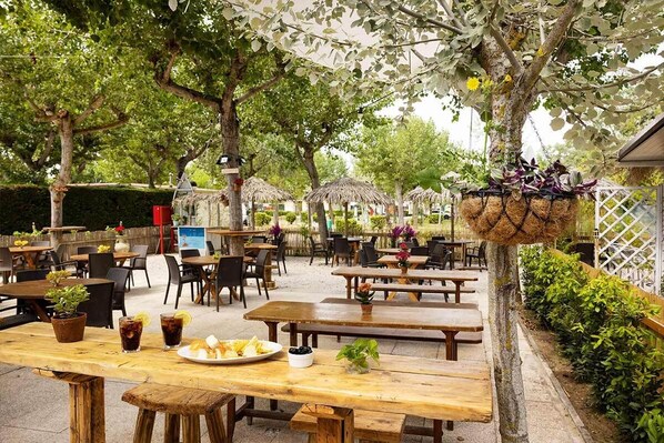 Un área de patio al aire libre con mesas y bancos de madera, rodeada de árboles y sombrillas de paja.