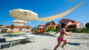 Une plage de sable avec des parasols, un espace piscine et des bâtiments colorés en arrière-plan.