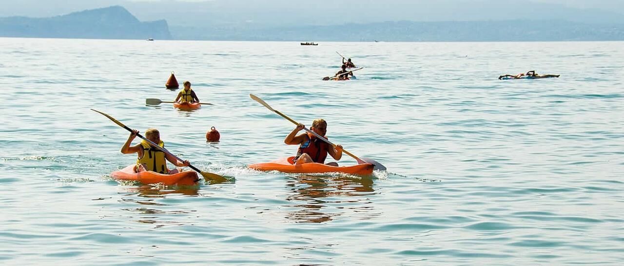 Menschen paddeln mit dem Kajak auf ruhigem Wasser, im Hintergrund erheben sich Berge.