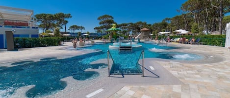 Ein Poolbereich mit verschiedenen Wasserspielen, darunter eine zentrale Fontäne und umliegende Pools, umgeben von Liegestühlen und Sonnenschirmen.