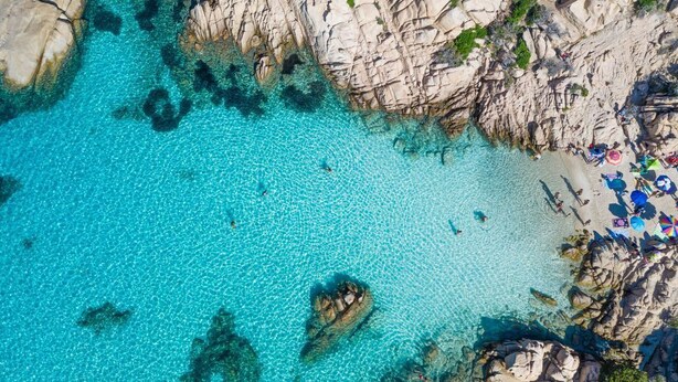 Vue aérienne d’une plage aux eaux turquoise et limpides, avec des formations rocheuses et des personnes se prélassant au soleil.